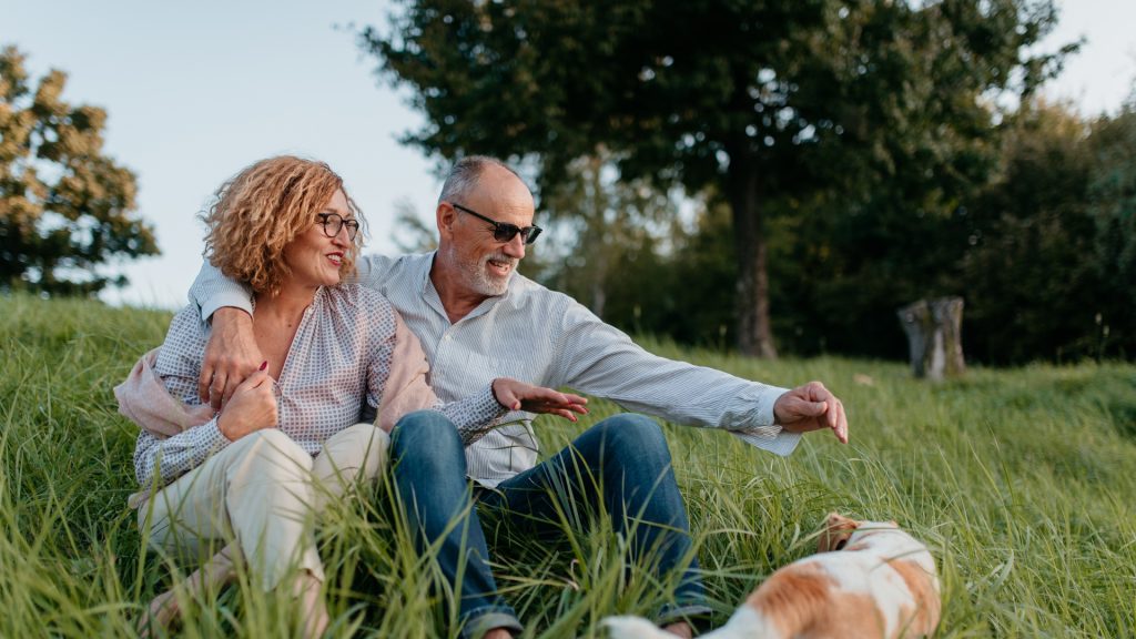 Two people sitting on a grassy hill with a dog. There is a tree and a view of the sky in the background.