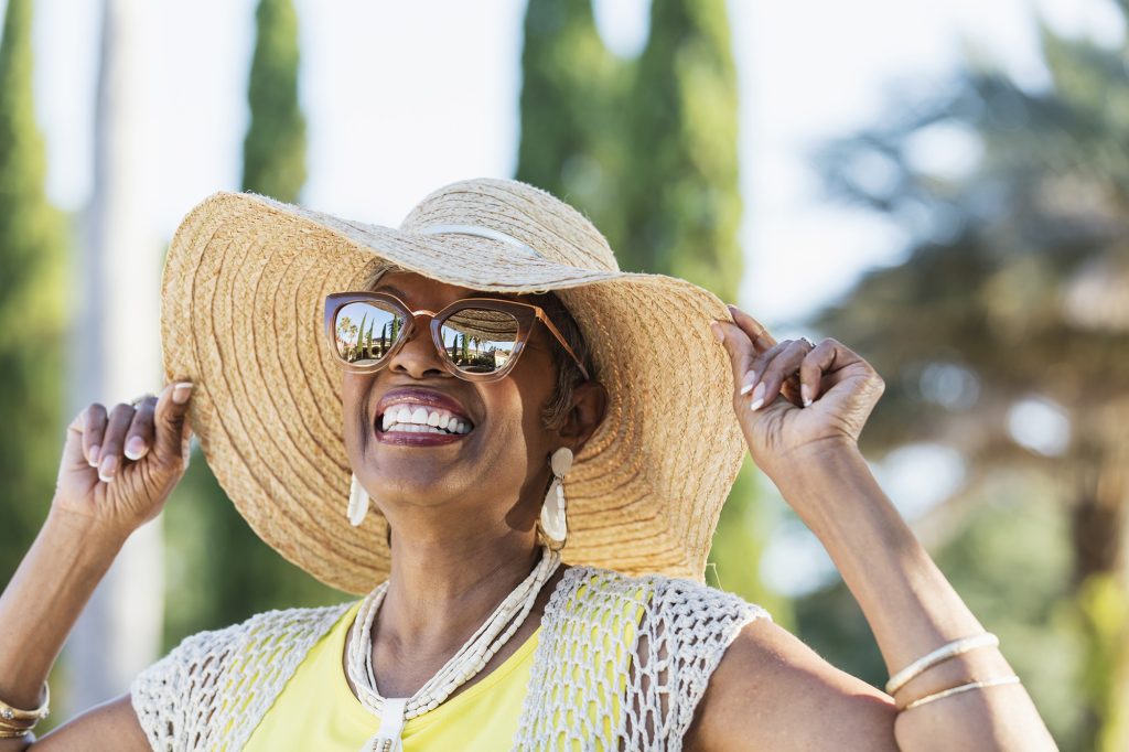 Someone wearing a sun hat and sun glasses. She is smiling and behind her are trees and a view of the sky.
