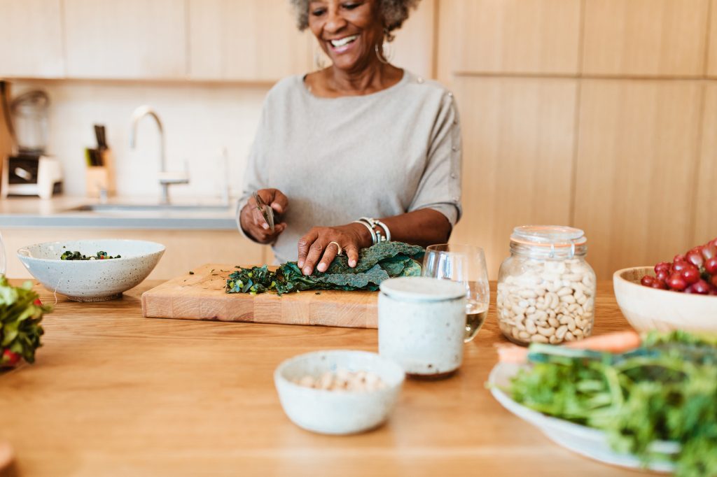 Someone cooking in kitchen and smiling. There are ingredients on the table and she is cutting kale with a knife.