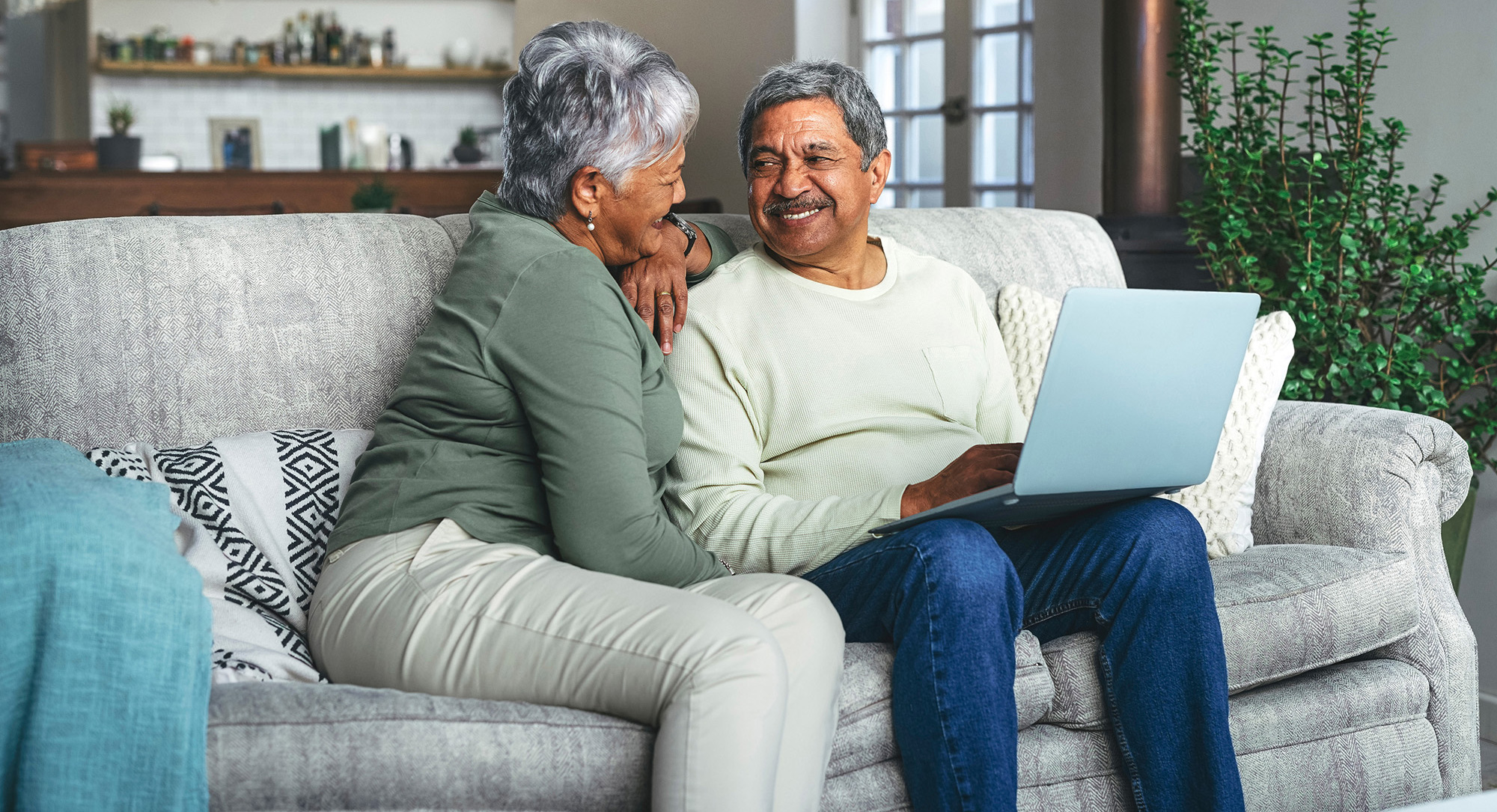 Two active adults sitting on a couch with a computer. The are looking at eachother and smiling lovingly.